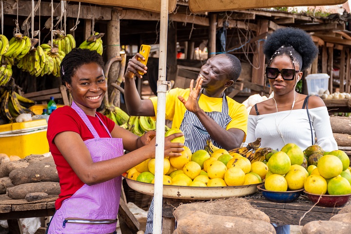 Local african market scene with happy traders selling, one using his phone to make video call.